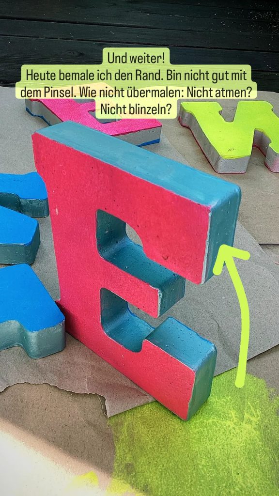 Ein sehr großer Betonbuchstabe E steht aufrecht zwischen anderen liegenden Betonbuchstaben. Er ist pink bemalt und an den Rändern blausilber.