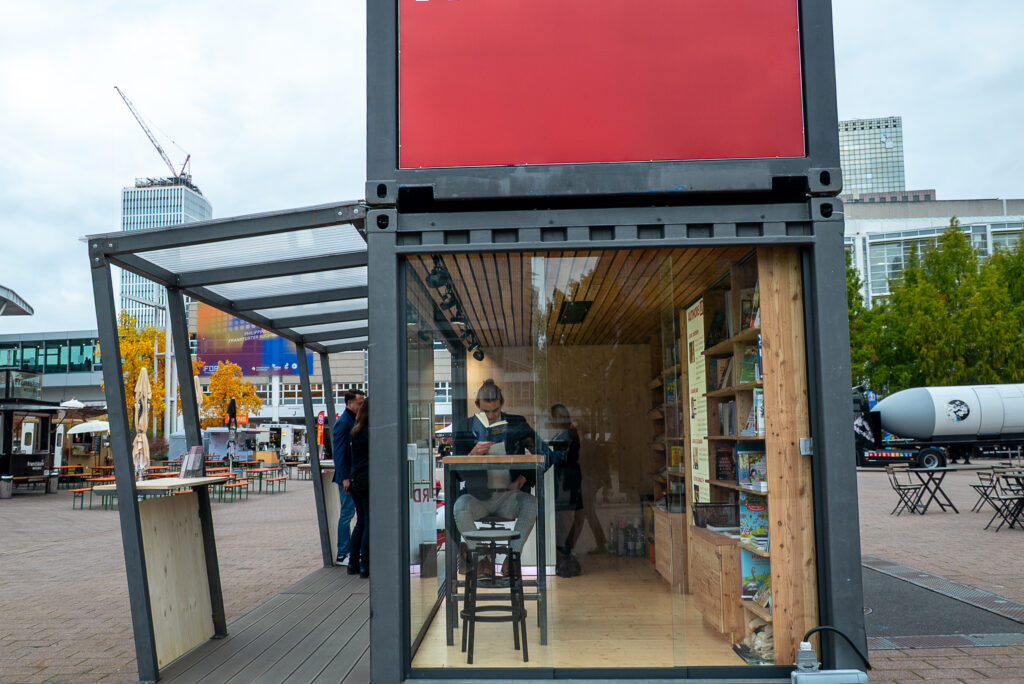 Auf dem Hof der Buchmesse steht ein Container mit durchsichtigen Wänden. An einem Bistrotisch sitzt ein Mensch und liest in einem Buch.