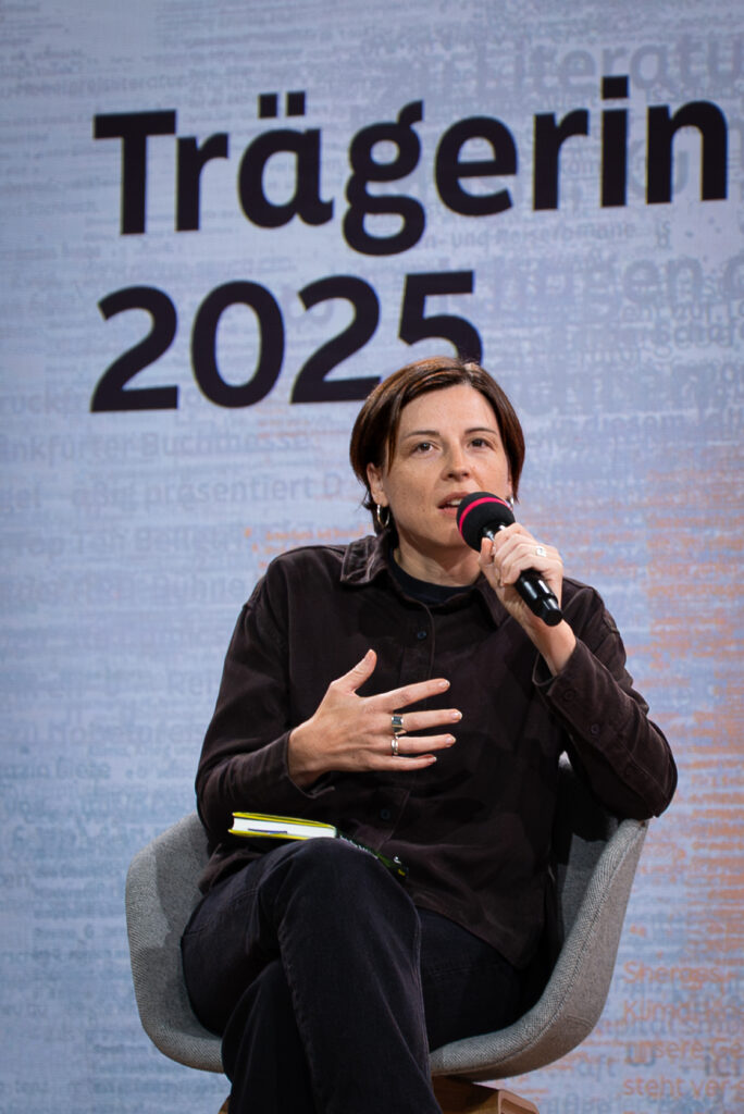 Dorothee Elmiger spricht gestikulierend in ein Mikro. Hinter ihr steht "Trägerin 2025" als Ausschnitt des Bildschirms