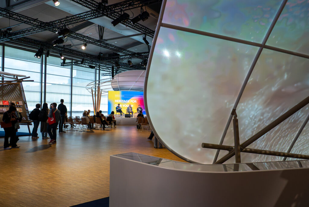 Blick vorbei am zentralen Screen auf die Bühne. Dort findet ein Panel statt zu "Heritage for our youth".