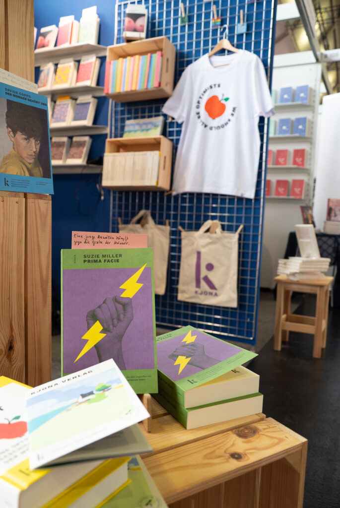 Sehr individuell und hübsch gestalteter Verlagsstand von Kjona mit Holzkiste, kleinen Bücherstapeln und Merch.
