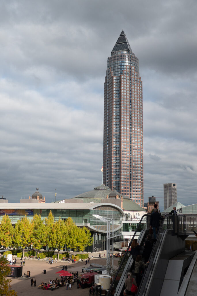 Im Hintergrund der Messeturm am Frankfurter Messegelände. Im Vordergrund fahren Besucher*innen mit der Rolltreppe in Halle 3.1.