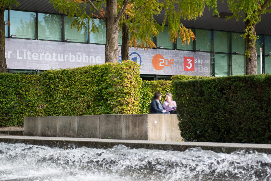 Gebäude der Literaturbühne von ARD, ZDF und 3sat im Hintergrund. Im Vordergrund ein Brunnen und Messebesucherinnen, die sich unterhalten.