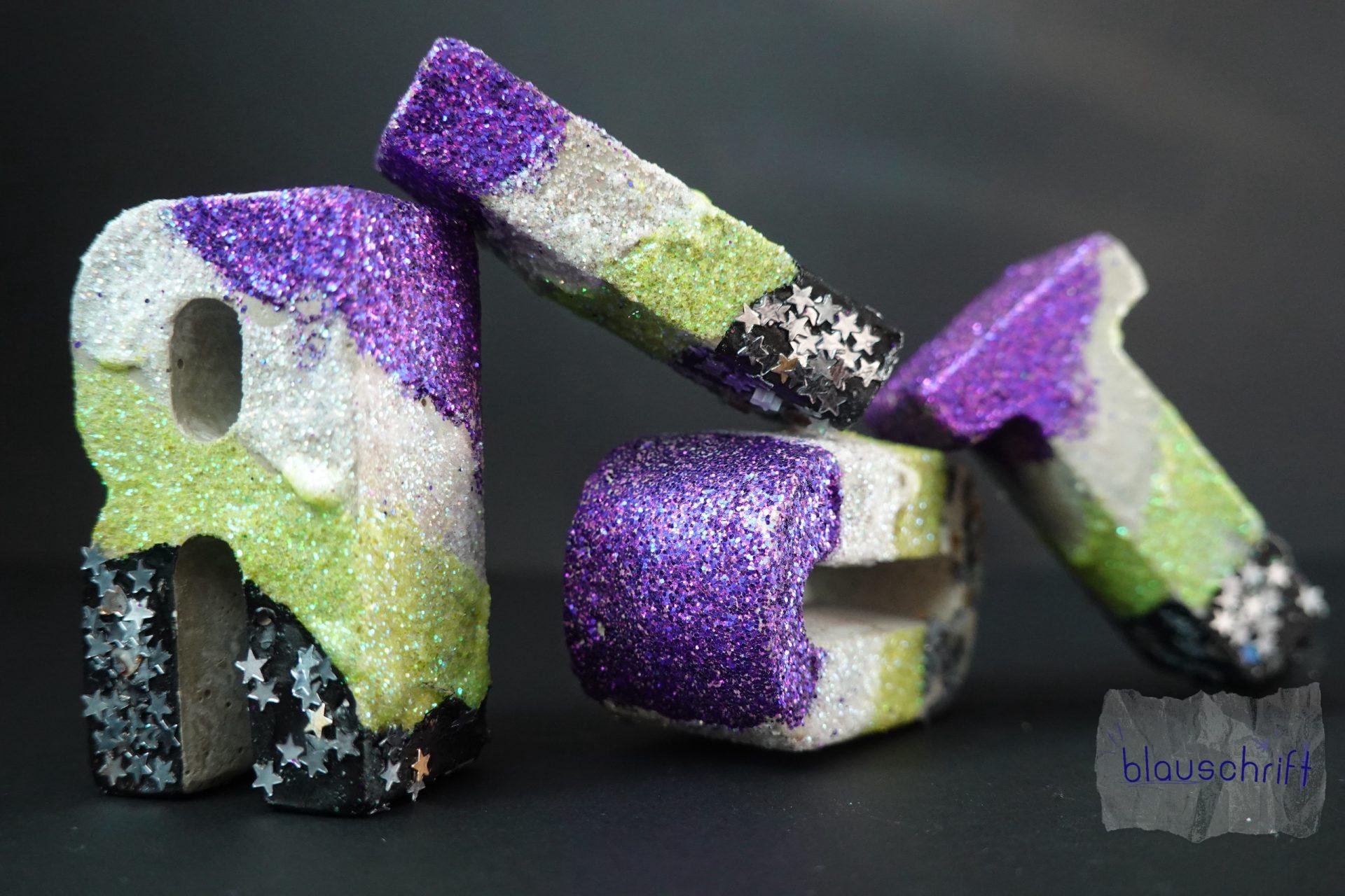 Betonbuchstaben lehnen aneinander: R I O T. Sie sind alle in den nonbinary Farben und glitzern.