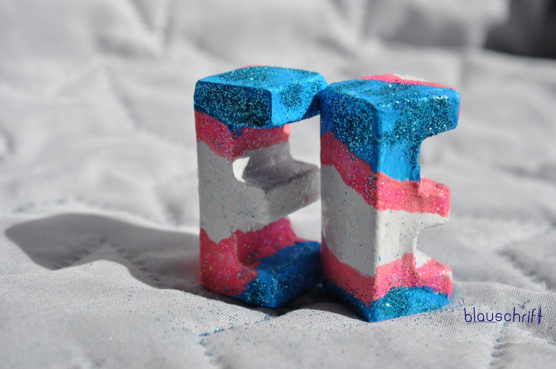 Zwei Betonbuchstaben E. Sie sind in trans pride Farben und glitzern.