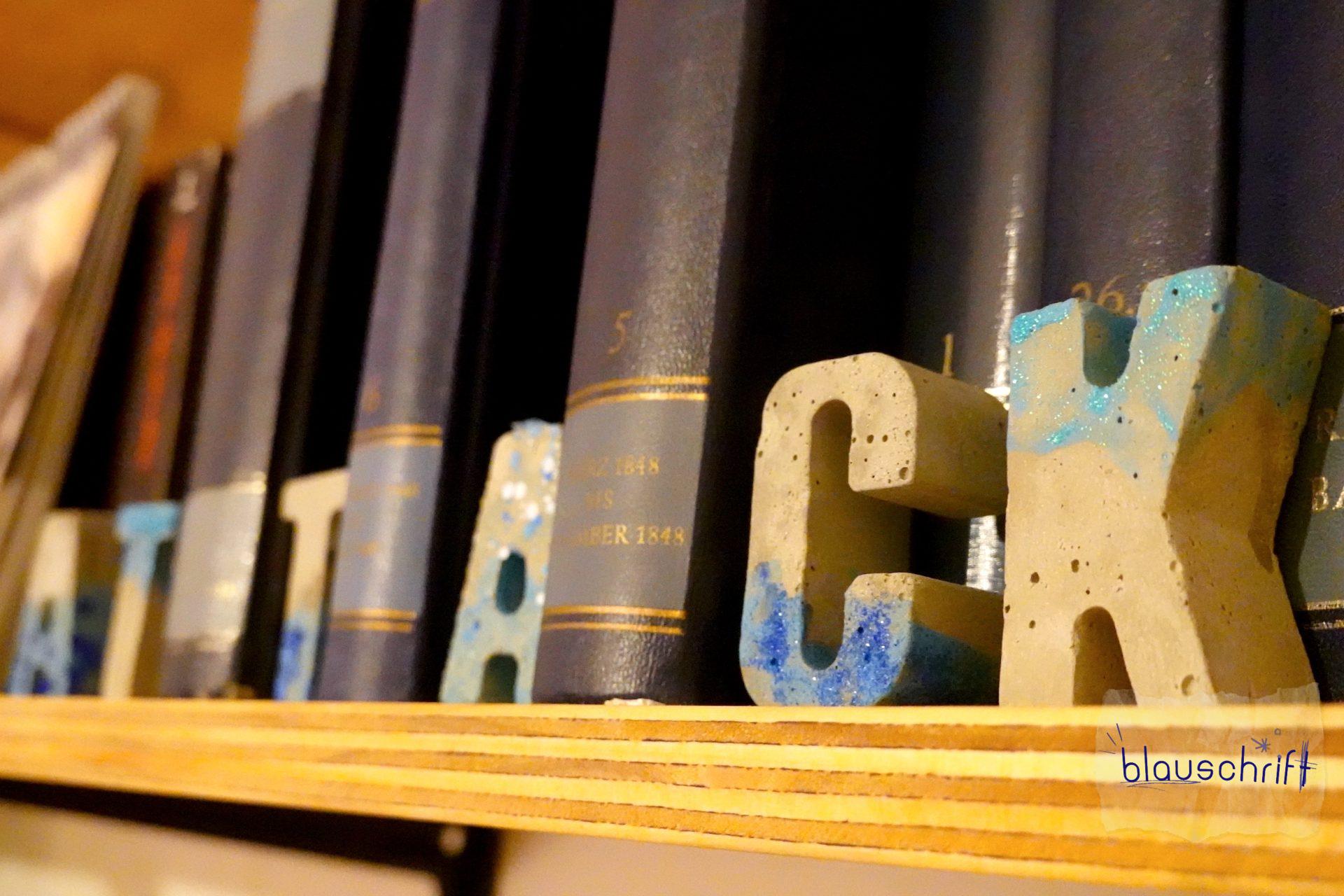 Kleine Betonbuchstaben stehen im Bücherregal an jedes zweite Buch gelehnt.