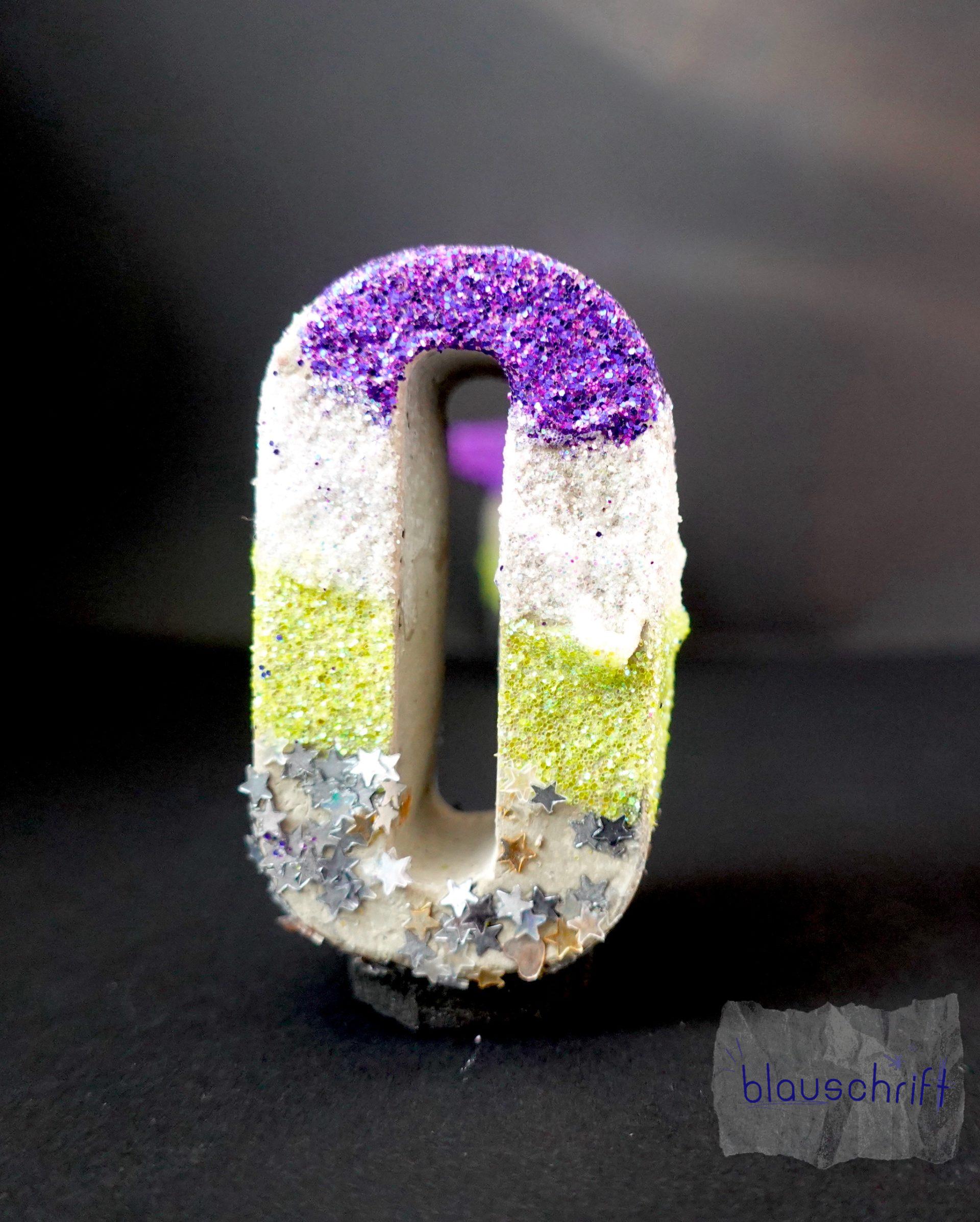 Kleiner Betonbuchstabe O. Er ist lila, weiß, gelb, schwarz und glitzert. Durch das O hindurch sieht man leicht das T.