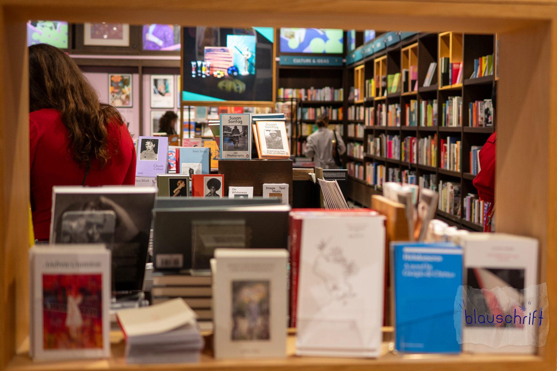 Durch ein Rgeal wie ein Guckfenster in den Buchladen fotografiert. Mehrere personen stöbern.