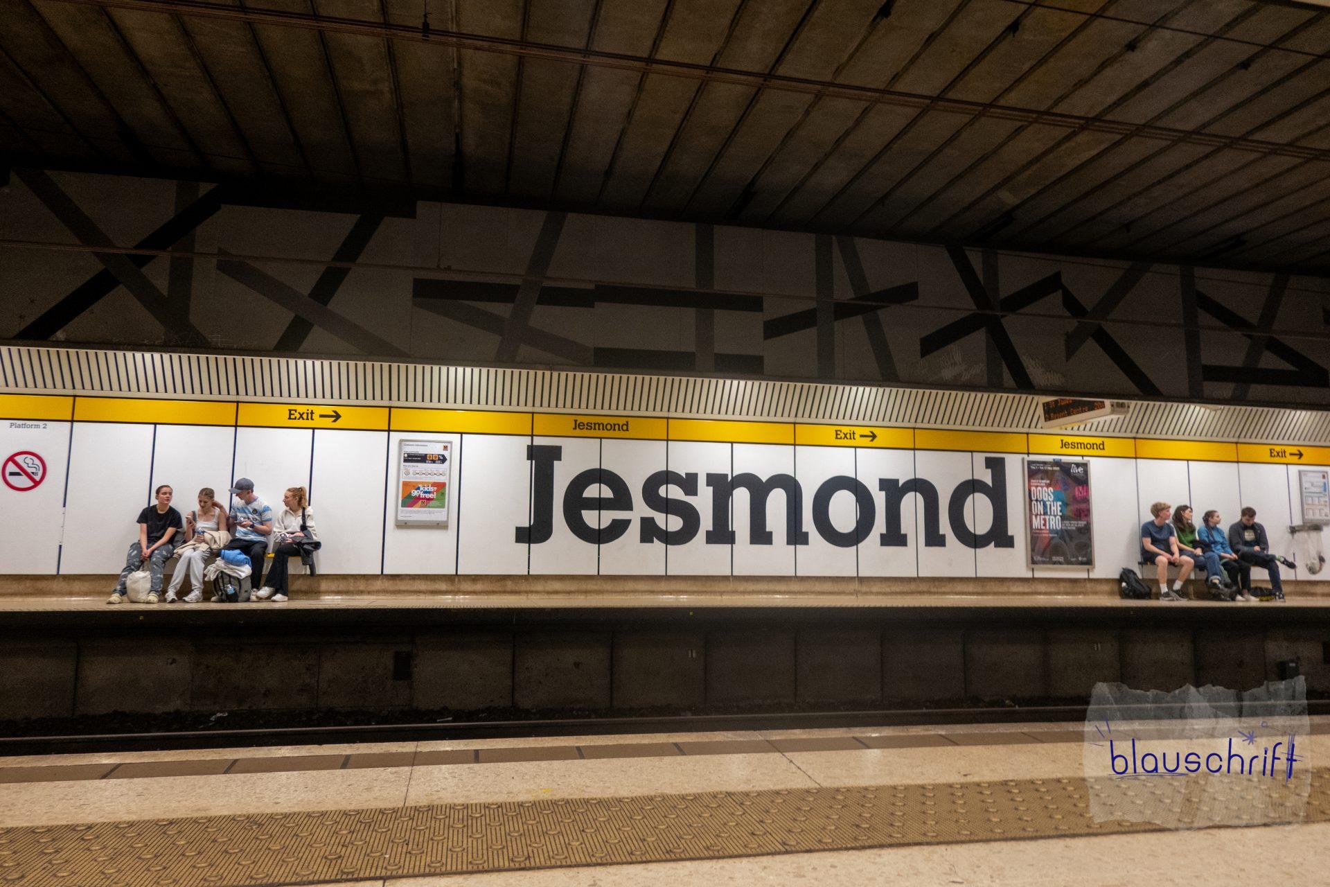 Metrohaltestelle "Jesmond" in Newcastle. Großer Schriftzug. Links und rechts davon sitzen Jugendliche auf Bänken und warten.
