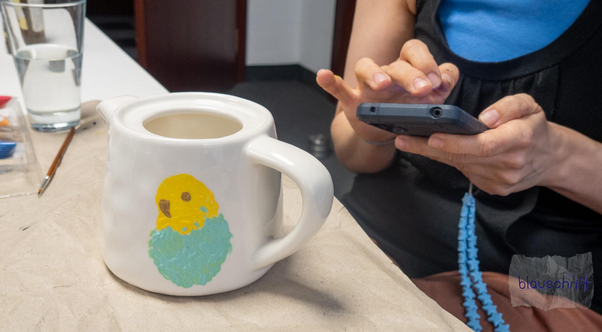 Alex Hände am Handy, vor ihr der Teekanne mit der Grundform vom drauf gemalten Wellensittich.