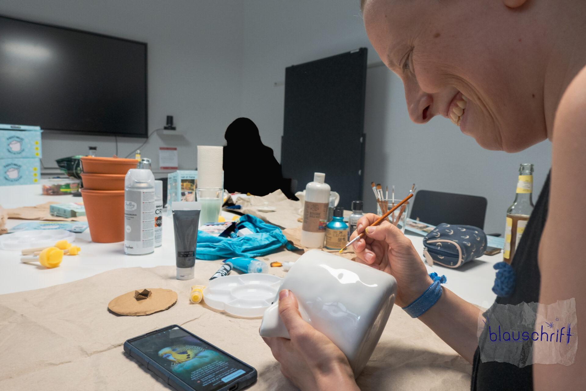Alex bemalt lachend ihre Teekanne. Sie schaut dabei auf ein Wellensittichbild auf dem Handy. Der Tisch ist voller Malutensilien.