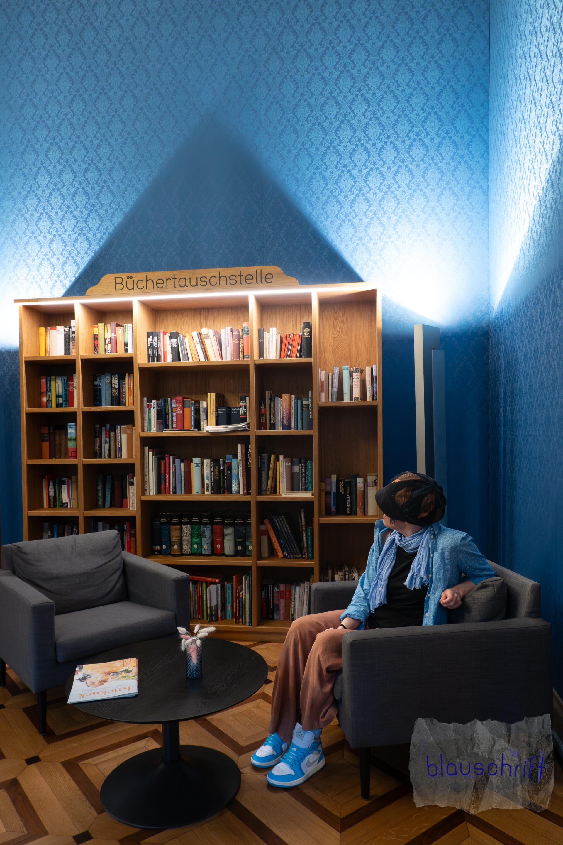 Alex sitzt in einem Sessel vor einem großen Büchertauschregal. Die Tapete ist so blau wie ihr Hemd.